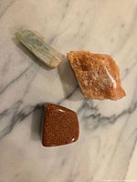 Three stones on marble surface: blue-green shard, orange calcite, brown tumbled stone