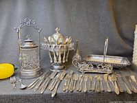 Group shot of all silver plated items including pickle caster, sugar spooner with spoons, goblets, bread basket, tray, butter spreaders