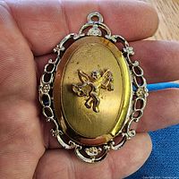 Front view showing gold-tone cherub motif, oval center, decorative border, bail
