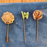 Three stick pins laid out on blue cloth: flower cabochon, enamel butterfly, rose