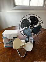Canadian Tire metal desk fan with four black blades beside Caframo cream plastic fan and box