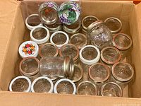 Box containing assorted small glass canning jars with various metal lids and sealed pack of rings