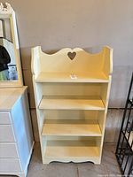Front view of cream painted wood bookshelf showing heart cutout, three shelves, scalloped top and base