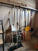 Wall-mounted assortment of long-handle garden tools, shovels, broom, roof rake, boot trays visible