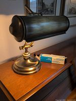 Side view of brushed antique brass piano lamp on wooden surface with spare bulb box beside it