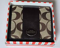 Front view of wallet in box showing canvas pattern, leather panel, brass logo plaque