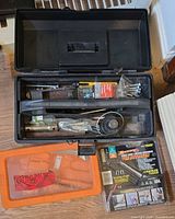 Open toolbox with assorted tools and hardware; Arrow staple gun package and orange bit set visible beside