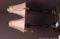 View of pair of metal lamps with light pink fabric shades, decorative beaded tassels, and dark metal bases with amber glass decor, placed on wooden surface.