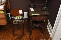 Wide shot of two side tables on hardwood floor; left is round wood top with metal legs, right is square carved wood table with ornate legs and lower shelf.