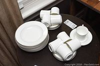 Group photo showing 8 white soup plates stacked, 8 matching cups and saucers arranged next to them on a dark round table.