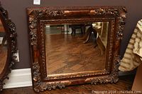 Full view of the rectangular vintage wall mirror with dark wood and gilded ornate carved frame resting on a wooden floor.