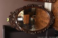 Full view of the vintage oval mirror with wooden carved frame showing floral and leaf motifs, on a wooden floor against a wall.