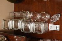 Four square glass canisters with lids and seals and four tea glasses laid out on a wood surface. Clear glass shown with square and cylindrical shapes.