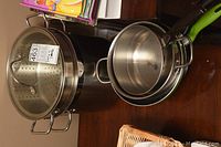 Photo showing a large stainless steel 12-quart Lagostina pot with a metal strainer insert and tempered glass lid, plus several smaller fry pans stacked together on table.
