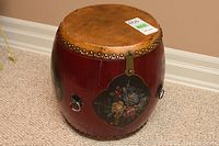 Front view of the drum-shaped wooden storage container showing the tan leather top with brass studs, reddish lacquer finish, and floral hand-painted panels on the side.
