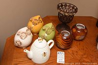 Group photo of 3 ceramic owls in white, yellow, green, white bee teapot, 2 brown woodgrain candle holders, and small metal basket holder on wood table.