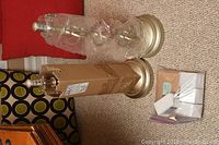 Pair of two Modern Lantern metal table lamps, one fully wrapped in plastic and the other partially wrapped with cardboard on the central body, placed on carpet with a red chair in the background