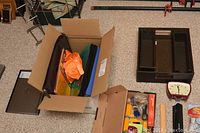 Image showing box containing colored-section laptop case or binder, brown laptop case, multiple items organized on carpet including dark brown office organizer, retro-look desk clock, black hole puncher, stationery items in box, and smaller desk clock.