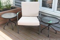 Outdoor metal garden chair with beige cushions and two round metal patio side tables with glass tops arranged on a wood deck outdoors.