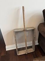 Full view of grain sieve leaning against wall showing frame, mesh, and handle