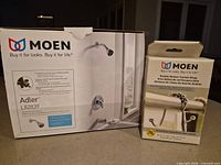 Boxed Moen Adler L82839 faucet set beside boxed Moen double shower curtain rings