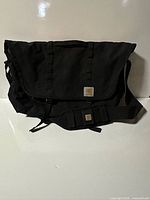 Front view of black Carhartt messenger bag showing flap, strap, and logo patch