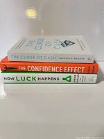 Three hardcover books stacked: The Curse of Cash, The Confidence Effect, How Luck Happens