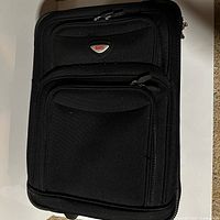 Front view showing black Roots carry-on with front zip pockets and brand badge