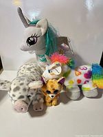Front view of four plush toys: grey unicorn, rainbow hearts unicorn, Ty cat, spotted pig