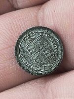 Front view of miniature shilling coin held between fingers, shows reverse shield design