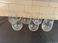 Six clear stemless wine glasses displayed upright on dark surface against tiled backsplash
