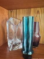 Three vases on wooden shelf: clear crystal Sails vase, turquoise cylinder vase, purple vase