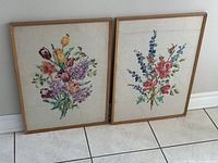 Both framed needlepoint pictures leaning against wall on floor tile