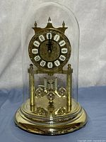 Frontal view of glass-domed brass-tone anniversary clock showing dial, pendulum and base