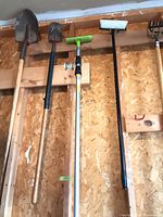 Five long-handled tools hanging on wall: shovel, cultivator rake, telescopic scrub brush, push broom