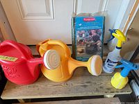 All items grouped on bench: two watering cans, two spray bottles, unopened Rubbermaid refuse bag