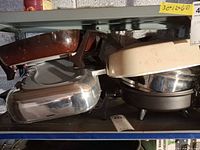 Stacked view of three skillets on shelf: brown enamel rectangular, polished stainless rectangular, cream round on base