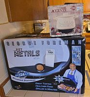 Front of George Foreman Hot Metals grill box with branding and specifications visible; Home Accents potpourri pot box stacked on top