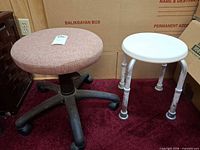 Swivel stool with pink cushioned seat next to white shower stool on red carpet, cardboard boxes backdrop