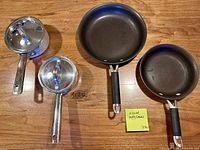 Four cookware pieces: two non-stick skillets, two stainless saucepans with lids displayed on wooden surface
