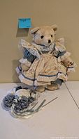 Beige teddy bear in blue floral lace dress standing, bonnet positioned in front