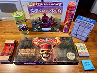 All seven boxed and tube games displayed on tabletop: Monopoly Pirates of the Caribbean, Small World, Melissa & Doug Suspend tube, Bowling Zombies tube, The New York Times Sudoku tin, Heavy Metal puzzle box, Places trivia card deck