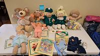 Table with five dressed Cabbage Patch Kids dolls, one headless body, plush teddy bear, outfits, papers, book