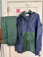 Front view of navy/green jacket with attached hood and matching green pants hung beside it