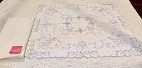 Full view of both linens: folded plain tablecloth and square eyelet overlay with scalloped border