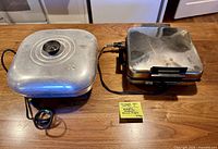 Waffle maker and skillet side by side on table with cords and lid visible