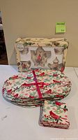 Box containing ceramic holiday cardinal tea set, stack of oval placemats, pile of matching cloth napkins