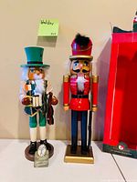 Both Kurt Adler wooden nutcracker figurines on bases; soldier in red coat and gentleman in green top hat; partial original box visible