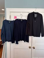 Front view of all three men’s tops hanging: slate blue v-neck, navy v-neck, black quarter-zip