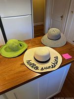 Three sun hats displayed on wooden island: green knit hat, beige straw 'hello sunshine' hat, black/white striped hat